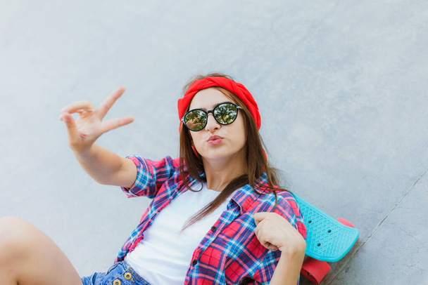 Young hipster girl in stylish mirrored glasses, shorts, red plaid shirt and bandage on her head lies with skateboard on concrete surface in skatepark - Photo, Image