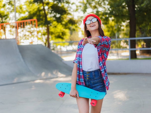 Cheerful young woman dressed in stylish clothes while holds skateboard in her hand in skatepark at bright sunny day - Photo, Image