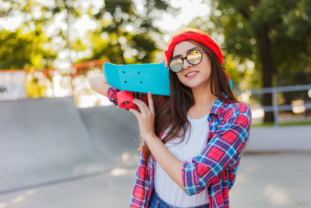 Radosna, emocjonalna młoda kobieta ubrana w stylowe ubrania i lustrzane okulary przeciwsłoneczne, trzymająca w ręku deskorolkę w skateparku w słoneczny dzień - Zdjęcie, obraz