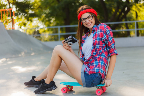 Śmieszna młoda kobieta w stylowych ubraniach siedzi na deskorolce i używa kamery retro w skateparku w pogodny słoneczny dzień. Koncepcja młodzieży - Zdjęcie, obraz