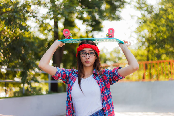 Zabawna młoda hipsterka ubrana w stylowe ubrania trzyma deskorolkę w ręku w skateparku w pogodny słoneczny dzień. Letni styl życia wizerunek modny ładna młoda dziewczyna  - Zdjęcie, obraz