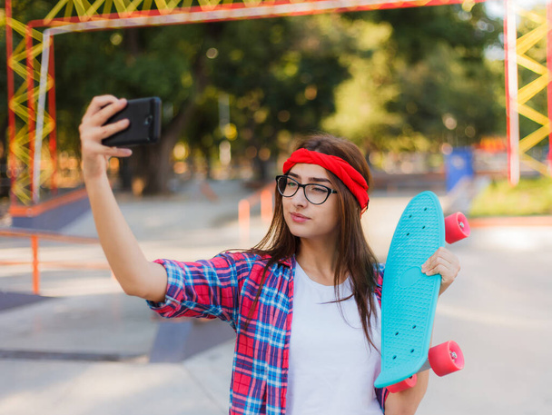 Funny young woman in glasses and red plaid shirt. Hipster girl n skatepark while makes  selfie with skateboard in skatepark at sunny bright day.  Summer fun - Photo, Image