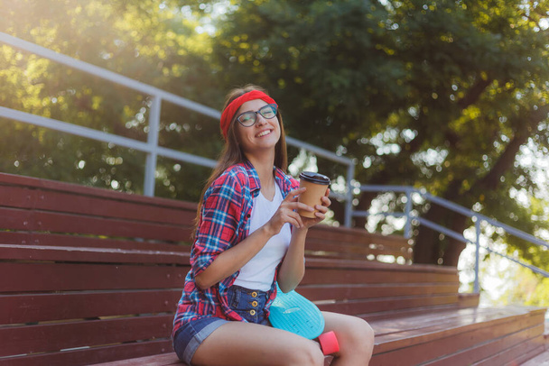 Young woman skater dressed in stylish clothes sits on the stands in a skatepark and drinks coffee on a bright sunny day - Fotoğraf, Görsel