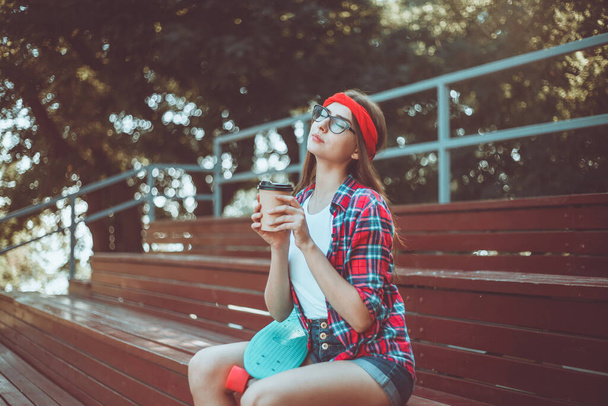 Mujer joven patinadora vestida con ropa elegante se sienta en las gradas en un parque de skate y bebe café en un día soleado brillante - Foto, imagen