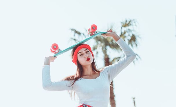 Portrait of stylish dressed young woman with skateboard outdoors on bright sunny day. Summer lifestyle image of trendy pretty young girl. - Photo, Image
