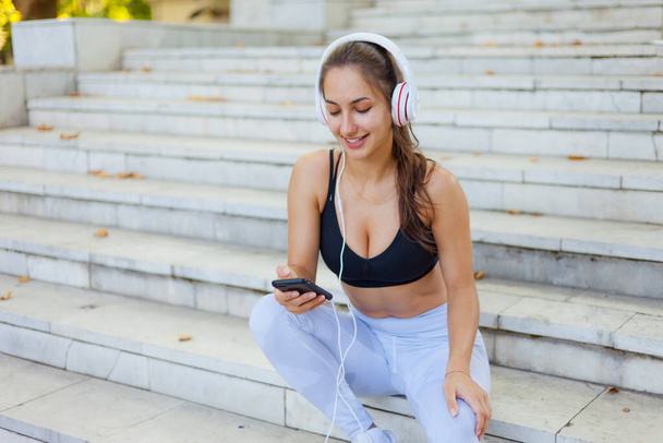 Concept of rest after training. Young happy sport woman in sportswear use smartphone and listens to music in headphones while sitting on stairs on bright sunny day - Photo, Image