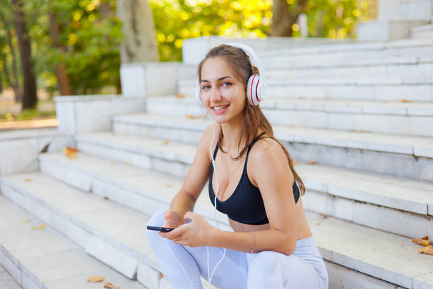 Concept of rest after training. Young sport woman in sportswear use smartphone and listens to music in headphones while sitting on stairs on bright sunny day - Photo, Image