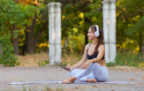 トレーニングと屋外レクリエーション。明るい晴れた日に公園のヨガマットに座っている間、若いスポーツ女性はヘッドフォンで音楽を聴く - 写真・画像