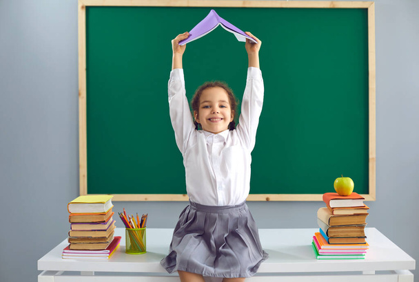 Back to school. Cheerful little girl sitting on desk and holding book above her head at classroom. Cute kid having fun at school - Foto, imagen