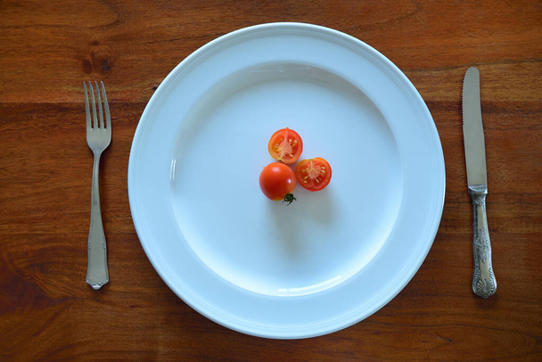 Ein großer weißer Teller von oben auf einer hölzernen Tischplatte mit Messer und Gabel und einer kleinen Anzahl roter Tomaten auf dem Teller - Foto, Bild