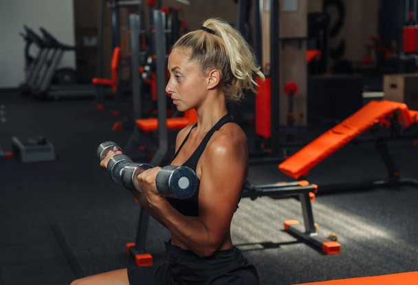Mladá fitness žena dělá činky pro biceps, zatímco sedí na lavičce v tělocvičně. Koncept školení s volnou hmotností. - Fotografie, Obrázek
