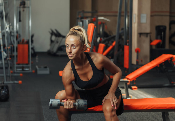 Young fitness woman doing concentrated doing dumbbell lifts for biceps one hand while sitting on a bench in the gym. Training concept with free weights. - Foto, Bild
