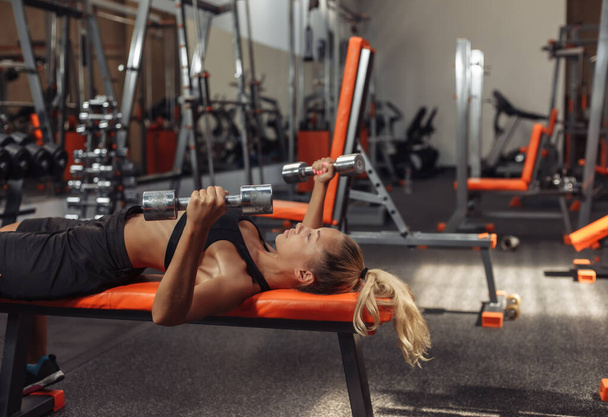 Slim fit woman in sportswear is practicing bench press with dumbbells while lying on a bench in the gym. Training concept with free weights. Functional training - Foto, immagini