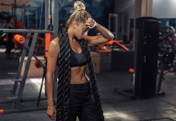 Mulher esportiva cansada em sportswear com corda de batalha em seu pescoço no ginásio. Treinamento funcional. Conceito de estilo de vida saudável
 - Foto, Imagem
