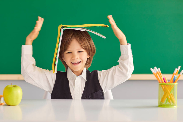 Back to school. Cheerful little boy hiding under book, playing silly game at desk in classroom. - Φωτογραφία, εικόνα