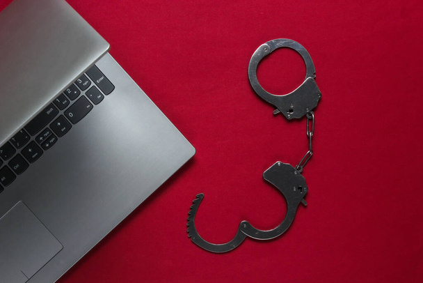 Кіберзлочинність, цифрова крадіжка в Інтернеті. Ноутбук зі сталевими наручниками на червоному тлі. Вид зверху
 - Фото, зображення