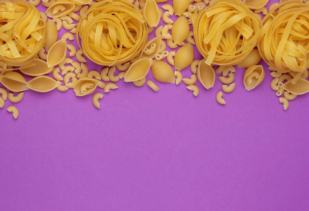 Verschillende soorten rauwe Italiaanse pasta op paarse achtergrond. Bovenaanzicht. Kopieerruimte - Foto, afbeelding