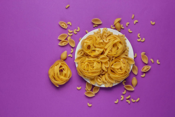 Különböző típusú nyers olasz tészta tányéron lila alapon. Felülnézet. - Fotó, kép