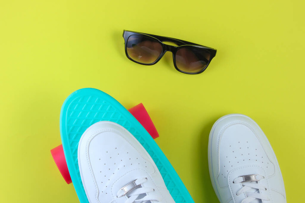 Λευκά αθλητικά παπούτσια σε πλαστικό mini cruiser board και γυαλιά ηλίου. Πράσινο φόντο. Καλοκαιρινή διασκέδαση. Άνω όψη - Φωτογραφία, εικόνα