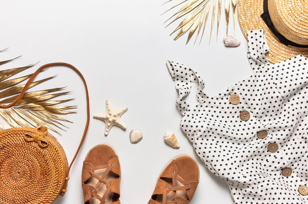 Summer women's white dress in black peas rattan woven bag brown sandals straw hat golden palm leaf shells starfish on light background. Flat lay top view. Women's beach fashion, travel vacation. - Photo, Image