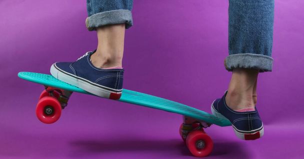Youth hipster concept. Female legs in jeans and sneakers on cruiser board in the studio on purple background. Summer fun - Foto, afbeelding
