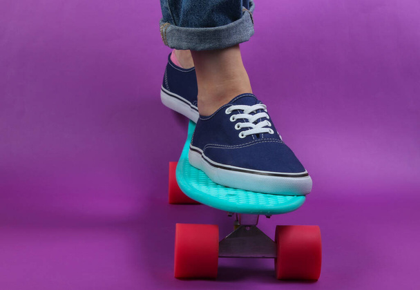Νεανική ιδέα χίπστερ. Γυναικεία πόδια σε τζιν και sneakers σε cruiser board στο studio σε μωβ φόντο. Καλοκαίρι διασκέδαση - Φωτογραφία, εικόνα