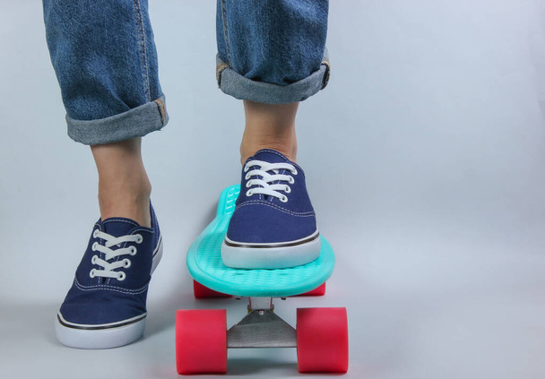 Νεανική ιδέα χίπστερ. Γυναικεία πόδια σε τζιν και sneakers σε cruiser board στο studio σε λευκό φόντο. Καλοκαίρι διασκέδαση - Φωτογραφία, εικόνα