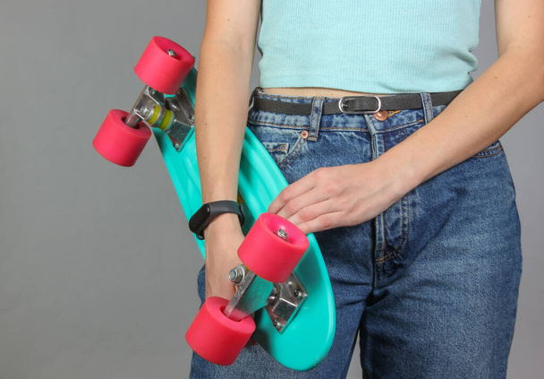 Young slim woman in jeans holds plastic skate board in her hands on a gray background. Youth hipster fashion. Crop photo - Photo, Image