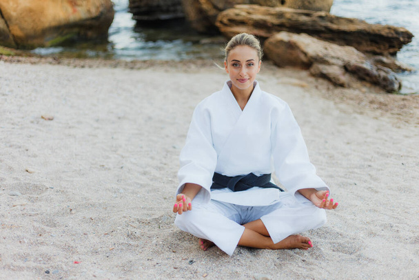 Young attractive female fighter in a white kimono with a black belt meditates on a wild beach with stones - Photo, Image