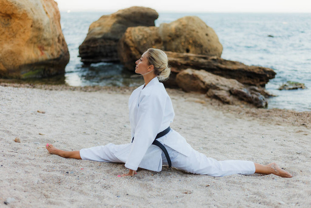 Mladý atraktivní karate žena v bílém kimonu s černým pásem sedí v motouzu na písku na divoké pláži s kameny - Fotografie, Obrázek