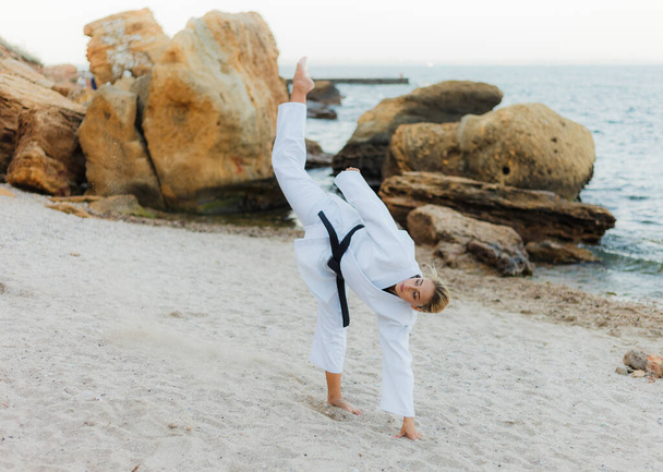 Jeune femme karaté attrayante en kimono blanc avec une ceinture noire trains coups de pied sur une plage sauvage avec des pierres - Photo, image