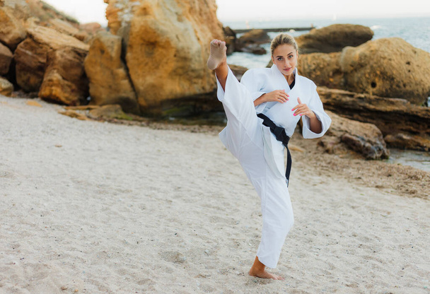Junge attraktive Karate-Frau im weißen Kimono mit schwarzem Gürtel trainiert Kicks an einem wilden Strand mit Steinen - Foto, Bild