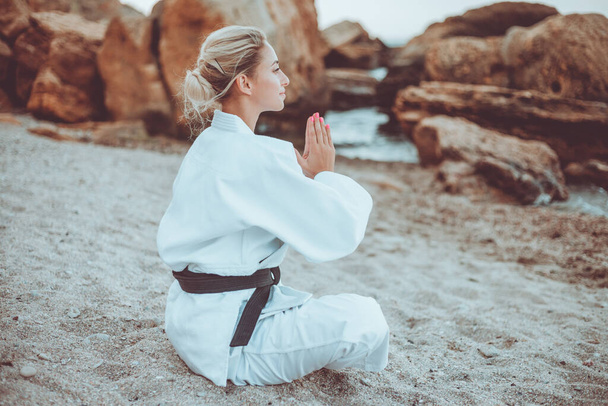 Atractiva artista marcial femenina en kimono blanco con cinturón negro se sienta en la arena y medita en una playa salvaje
 - Foto, imagen