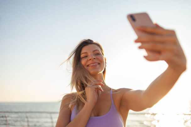 Selfie portrait of a young blonde woman dressed in sportswear on the beach at sunrise - Foto, Imagem