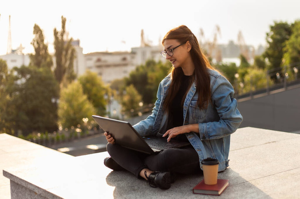 Jonge moderne studente in een spijkerjasje zittend en maakt gebruik van laptop outdoor. Afstandsonderwijs. Modern jeugdconcept. - Foto, afbeelding
