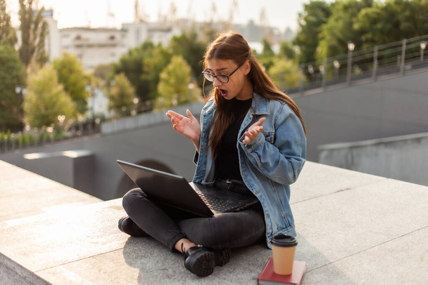 Meglepett modern diák nő farmerdzsekiben ül a lépcsőn és nézi a képernyős laptopot a szabadban. Távolságtanulás. Modern ifjúsági koncepció. - Fotó, kép