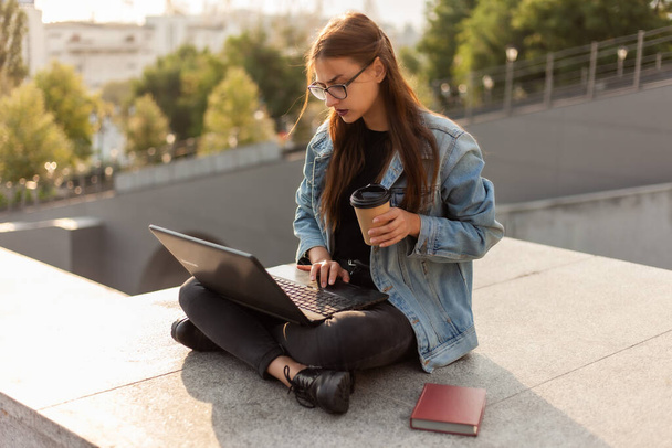 Young modern woman student in a denim jacket sitting on the stairs with laptop. Distance learning. Modern youth concept. - Photo, Image