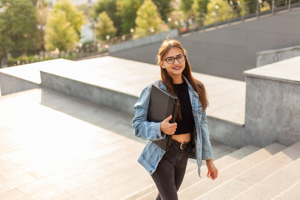 Jeune étudiante dans une veste en denim et des lunettes monte les escaliers avec un ordinateur portable dans ses mains dans la ville - Photo, image