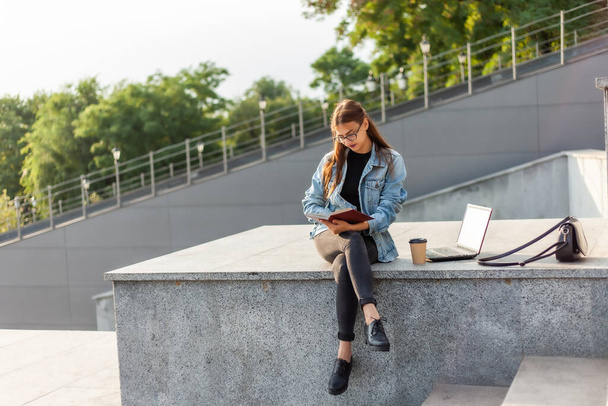 Young attractive student woman is reading a book while sitting on the stairs in the city. The concept of distance learning. Modern student - Photo, Image