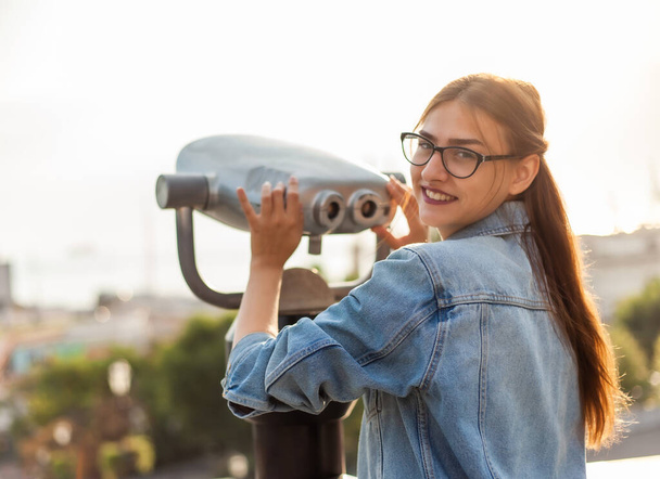 Jonge vrouw toerist in een spijkerjasje en bril kijkt in de stadsverrekijker in de stad. Reisconcept - Foto, afbeelding