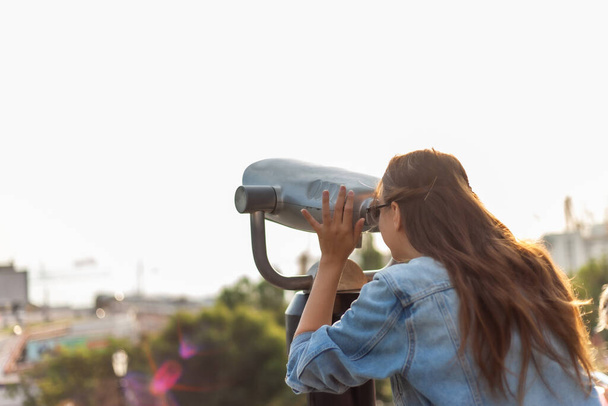 Mladá turistka v džínové bundě a brýlích se dívá do městského dalekohledu ve městě. Cestovní koncept - Fotografie, Obrázek
