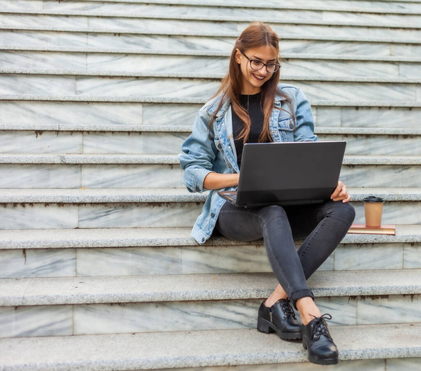 Kot ceketli genç modern bayan öğrenci merdivenlerde oturuyor ve dizüstü bilgisayarda video izliyor. Mesafe öğrenme. Modern gençlik kavramı. - Fotoğraf, Görsel