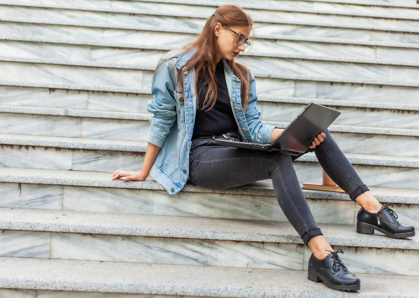 Jeune étudiante moderne dans une veste en denim assis sur les escaliers avec ordinateur portable. Je regarde la vidéo. Enseignement à distance. Concept de jeunesse moderne. - Photo, image