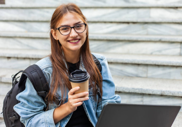Happy modern woman student in denim jacket and backpack sitting on the stairs with laptop outdoor. Drinks coffee. Distance learning. Modern youth concept. - 写真・画像