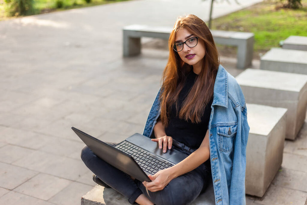 Młoda, nowoczesna studentka w dżinsowej kurtce, siedząca na schodach z laptopem. Uczenie się na odległość. Nowoczesna koncepcja młodzieży. - Zdjęcie, obraz