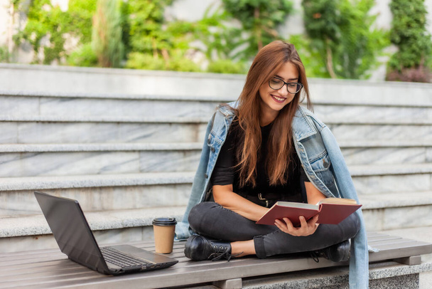 Modern öğrenciler. Mesafe öğrenme. Genç hevesli kadın dizüstü bilgisayarla bankta otururken kitap okuyor. - Fotoğraf, Görsel