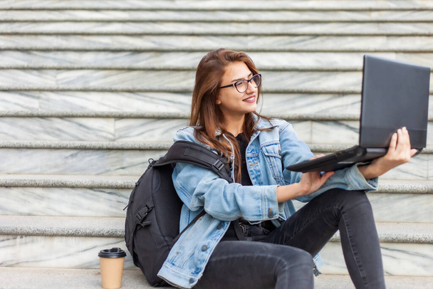 Junge, moderne Studentin in Jeansjacke und Brille sitzt mit Laptop auf der Treppe. Video ansehen. Fernstudium. Modernes Jugendkonzept. - Foto, Bild