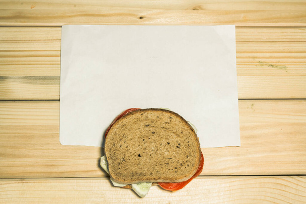 Egy vegetáriánus szendvicsre, ami néhány szelet paradicsomból és néhány szelet uborkából készült egy fehér papírdarab mellett, fából készült háttérrel. - Fotó, kép
