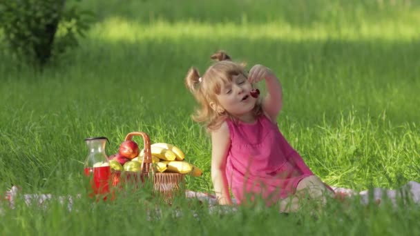 Week-end au pique-nique. Belle fille caucasienne enfant sur herbe verte prairie manger joyeux, cerise - Séquence, vidéo