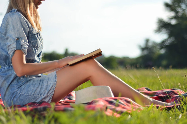 Girl reading book in nature on sunny summer day. Blonde attractive woman holds book on legs and wears jeans skirt. Girl sitting on blanket. Relaxation in nature concept. Close up, selective focus - Photo, Image